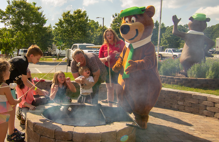 Yogi bear mascot roasting marshmallows over a fire pit with a family