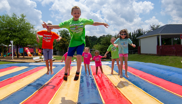 7 children jumping on inflatable jump pad with grass and sunny skies behind
